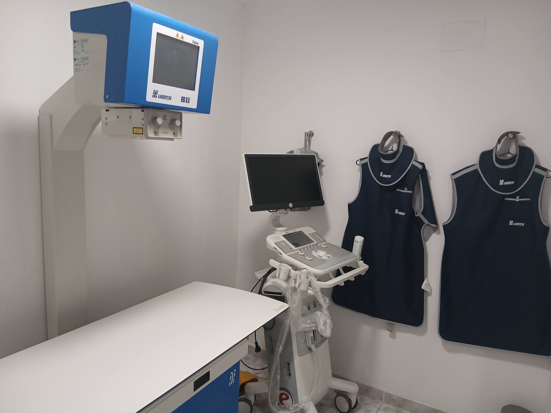 Una estación de rayos X médica con una camilla de exploración blanca, un monitor azul y un delantal de plomo colgado en la pared.