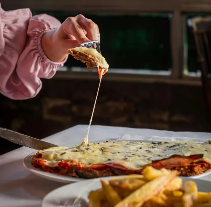 Persona sosteniendo con un tenedor una porción de milanesa con queso. Primer plano del plato acompañado de patatas fritas.