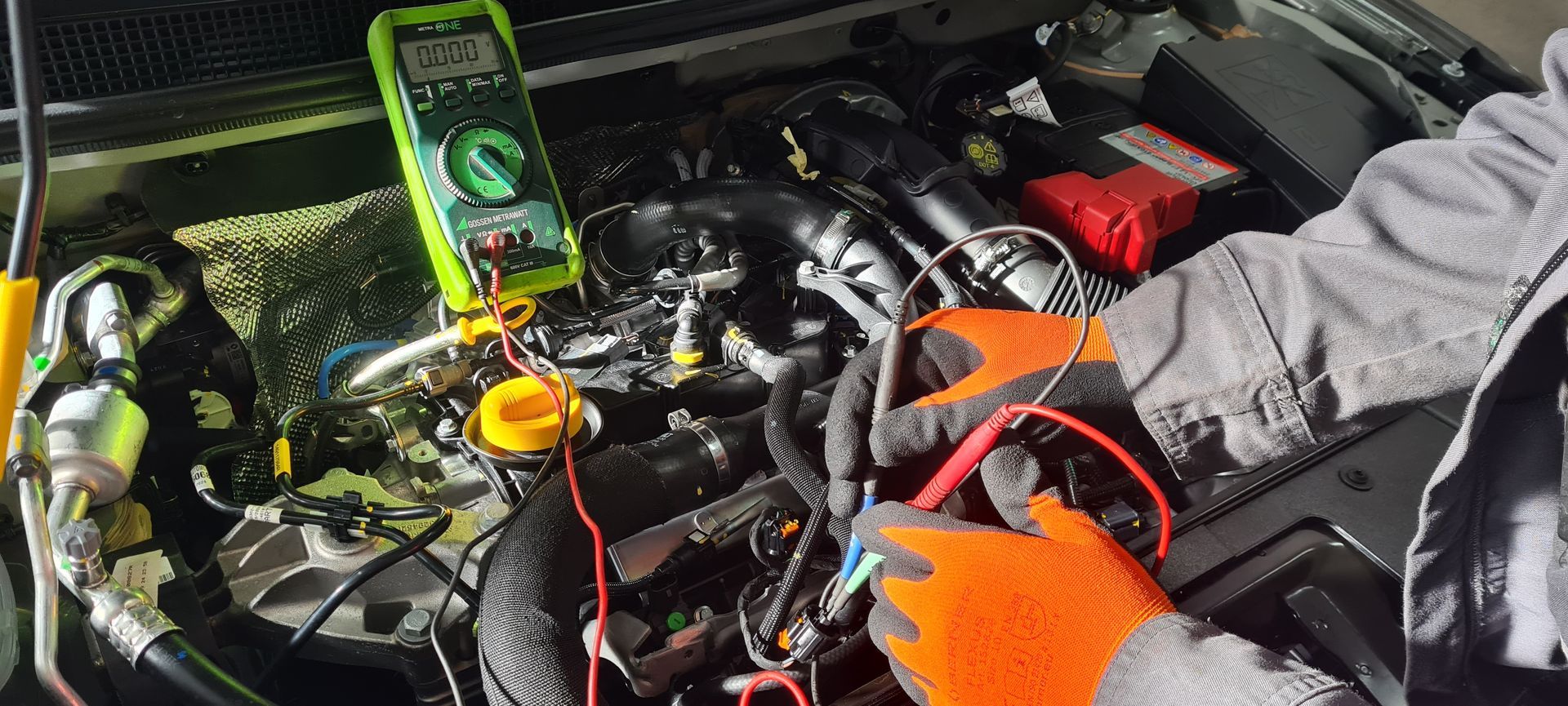 Eine Person mit orangefarbenen Handschuhen testet im Freien einen Automotor mit einem Multimeter.