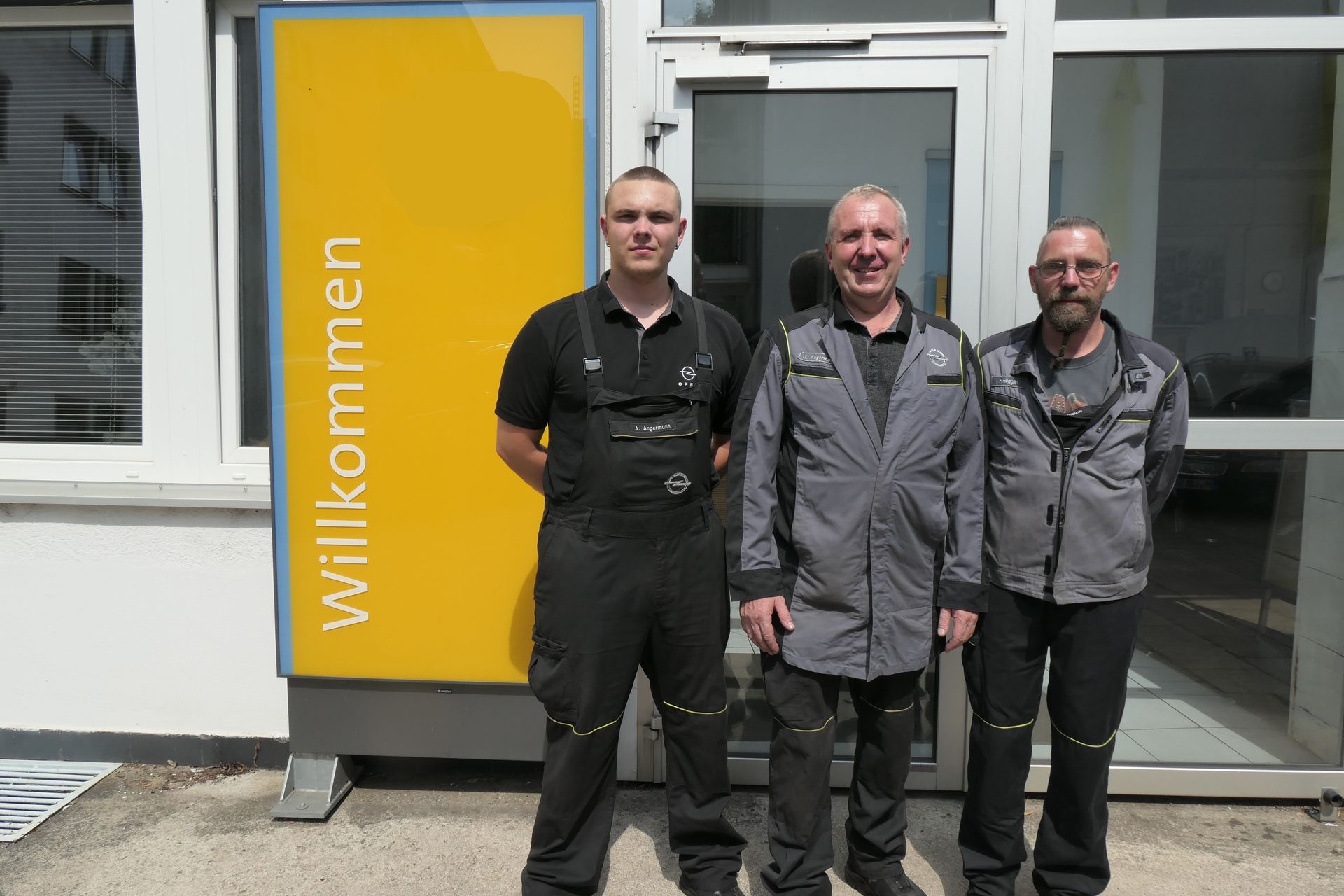 Drei Männer in Arbeitskleidung stehen vor einem Gebäudeeingang mit einem „Willkommen“-Schild.