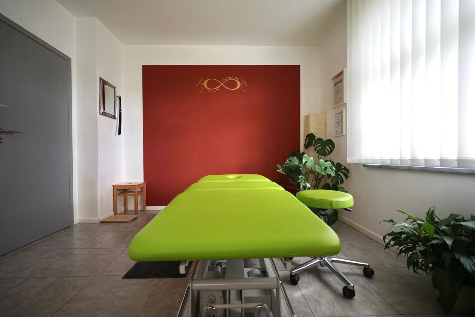 In einem Raum mit einer roten Wand steht eine grüne Massageliege.