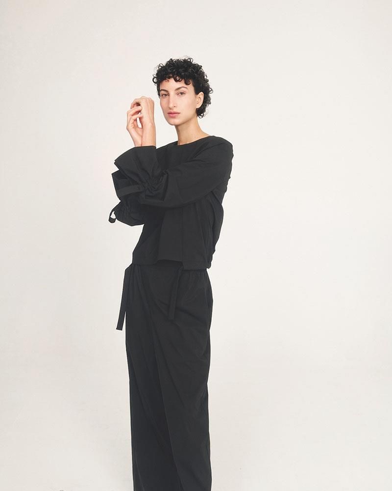 Eine Frau in einem schwarzen Overall steht mit verschränkten Armen da.