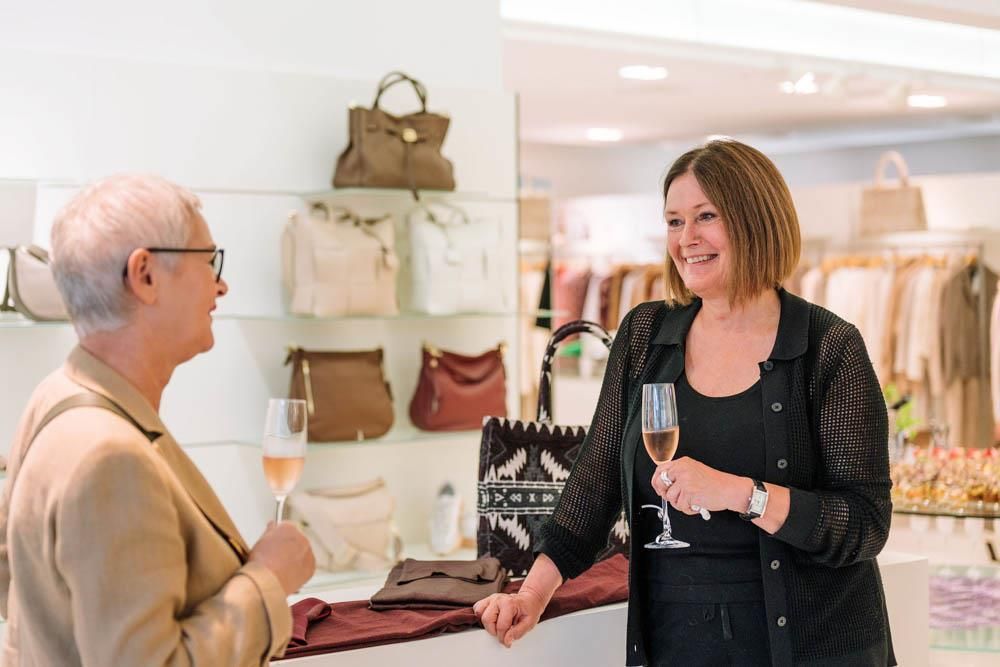 Eine Frau hält ein Glas Wein in der Hand , während sie in einem Geschäft mit einer anderen Frau spricht.