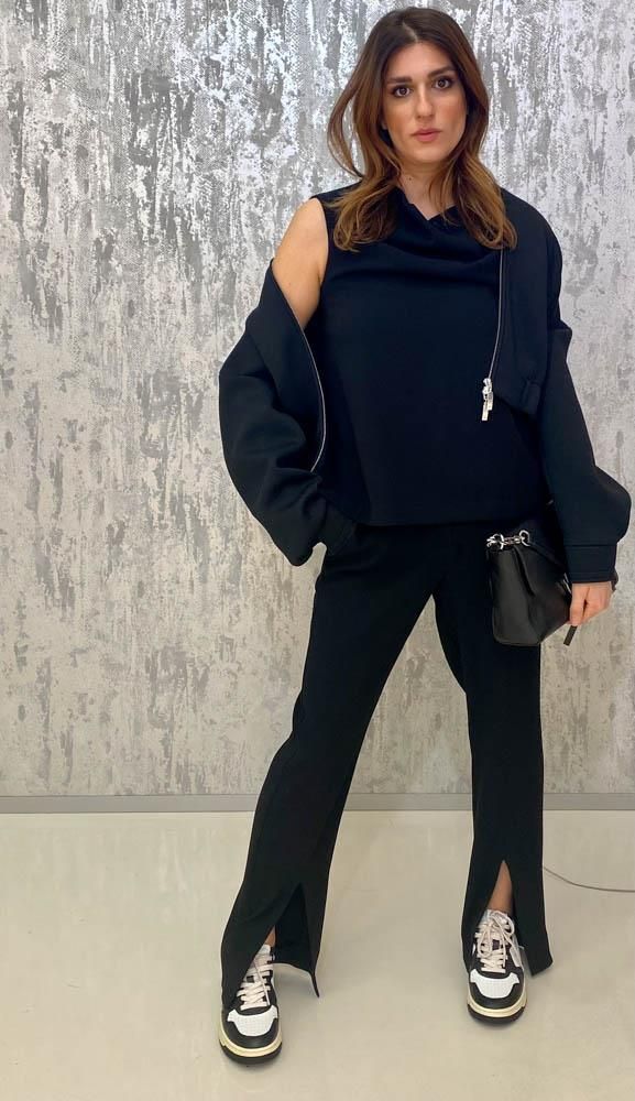 Eine Frau steht mit einer schwarzen Jacke und einer schwarzen Hose vor einer Wand.