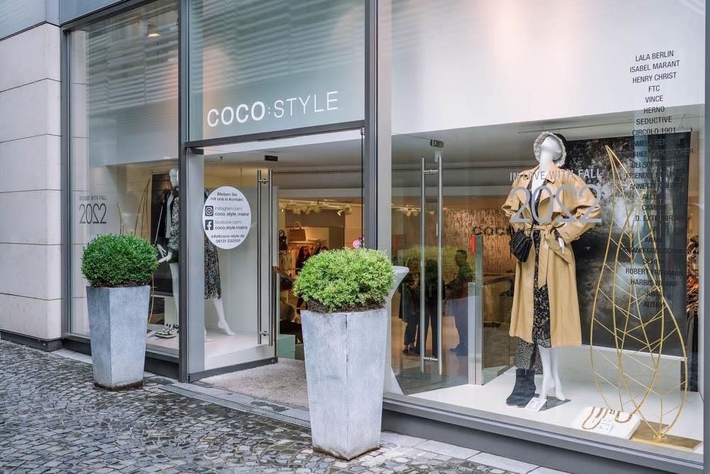 Eine Ladenfront mit einer Schaufensterpuppe im Fenster und Topfpflanzen davor.