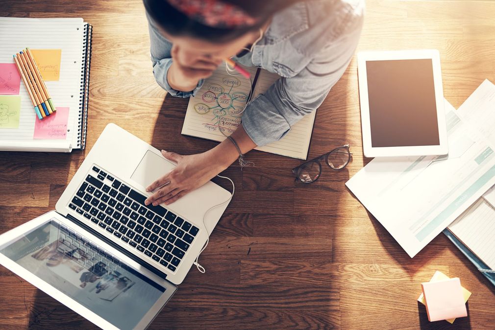Une personne travaille sur un ordinateur portable à un bureau en bois, entourée de papiers, d'un carnet, de lunettes et d'une tasse de café.