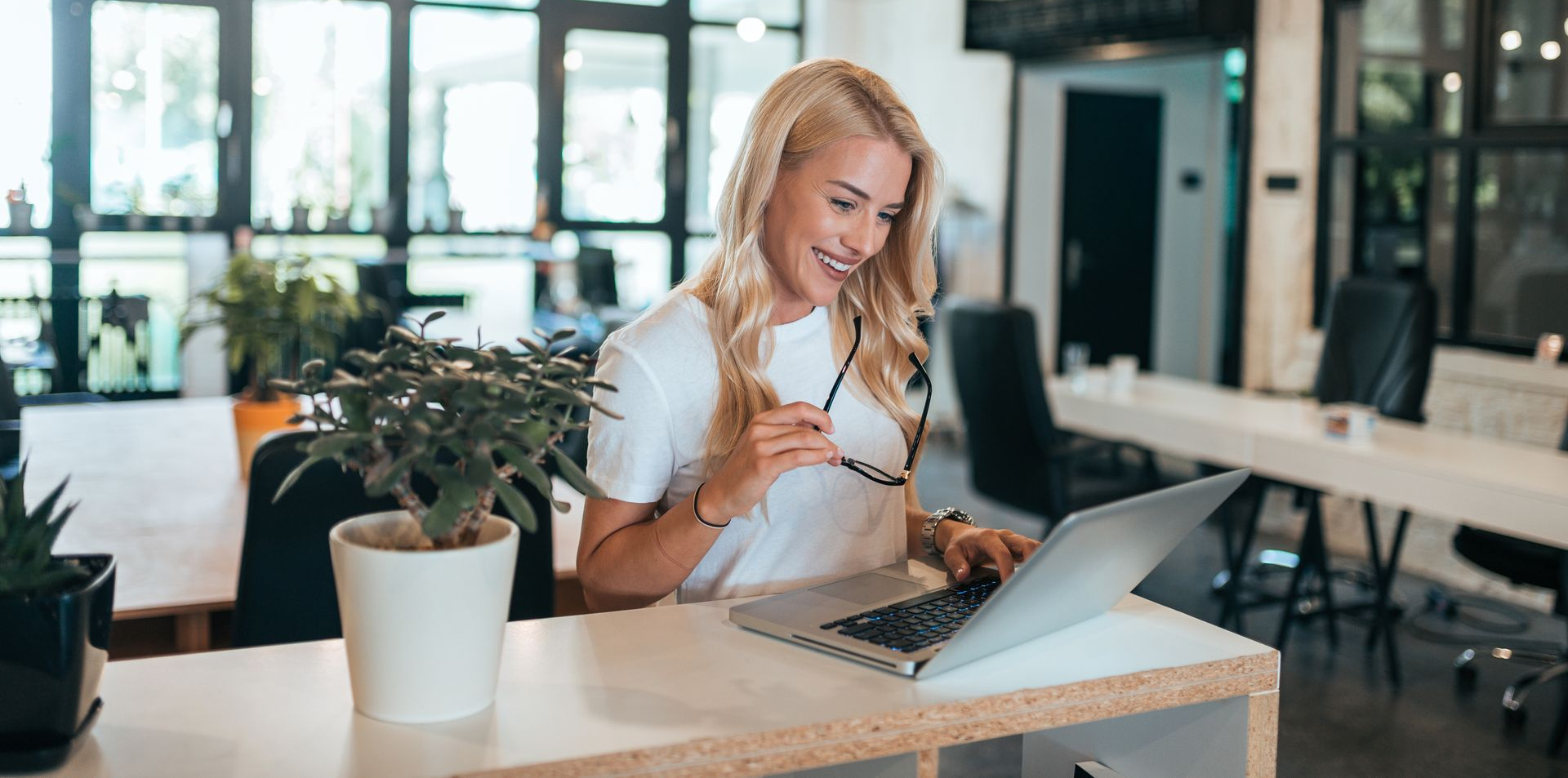Une femme sourit devant son ordinateur portable dans un bureau moderne et lumineux, à côté d'une plante en pot.