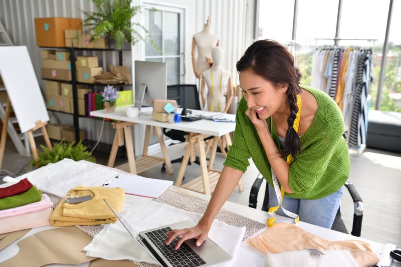 Femme dans un studio lumineux utilisant un ordinateur portable à une table de travail entourée de tissu, d'outils de couture et de matériel de conception.