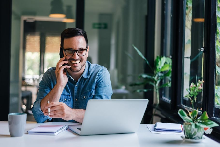 Une personne, souriante, est assise à son bureau, un ordinateur portable devant, au téléphone, dans un bureau moderne.