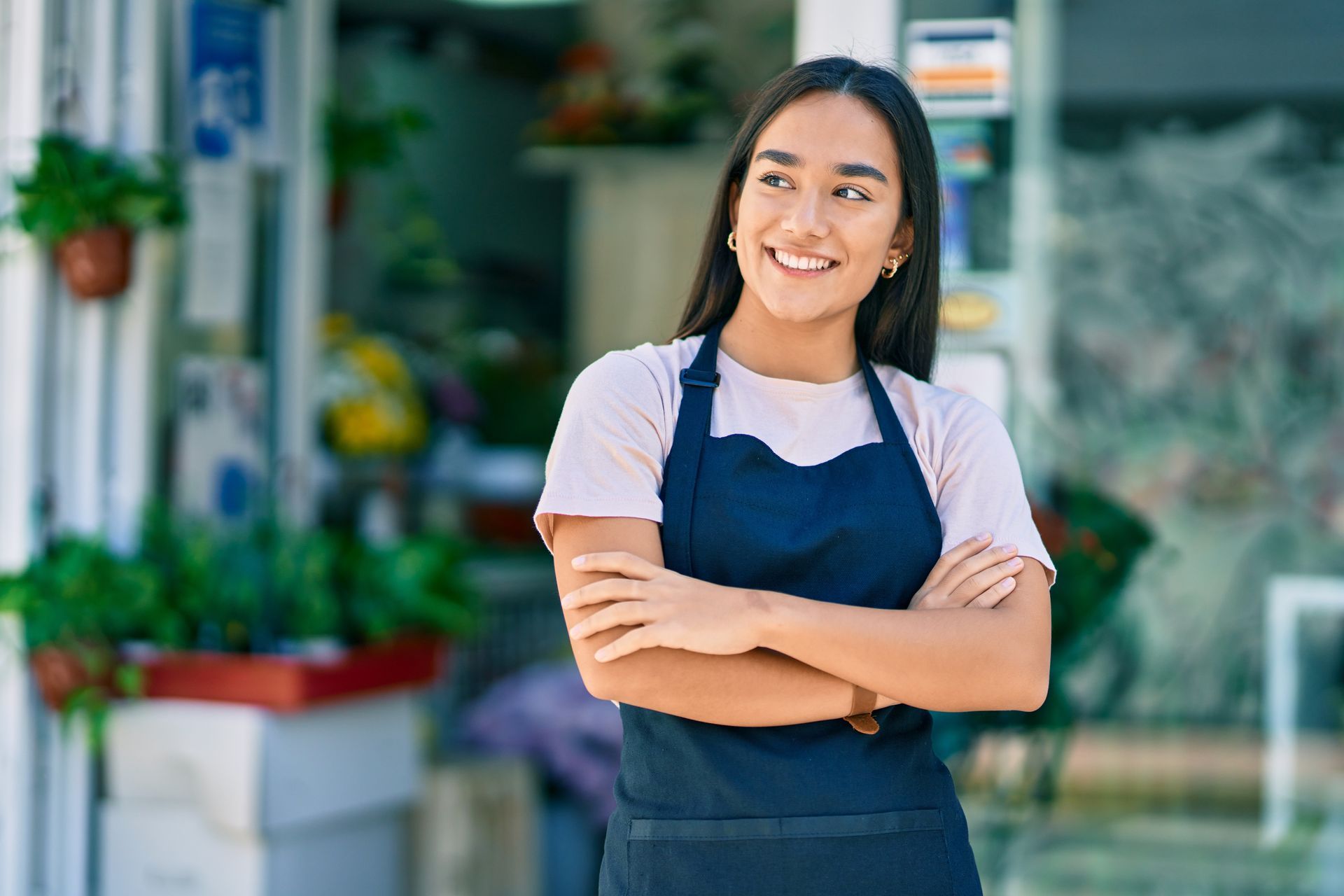 Employée de magasin souriante en tablier bleu marine, les bras croisés, devant une vitrine