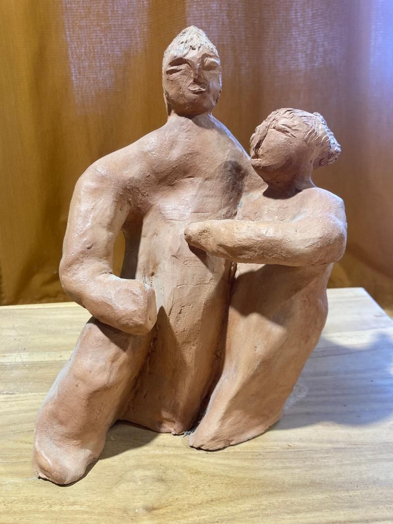 Deux figurines abstraites en argile assises côte à côte sur une table, devant un fond brun.