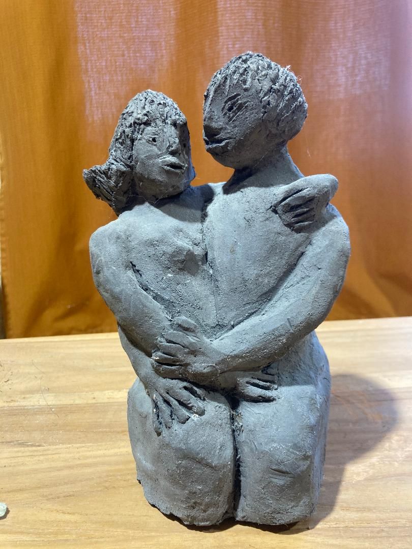 Sculpture grise de deux personnages enlacés sur une surface en bois, devant un rideau orange en arrière-plan.