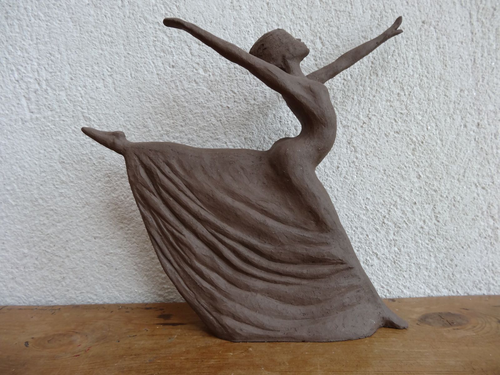 Sculpture abstraite brune représentant une danseuse aux bras levés, posée sur une surface en bois contre un mur texturé..