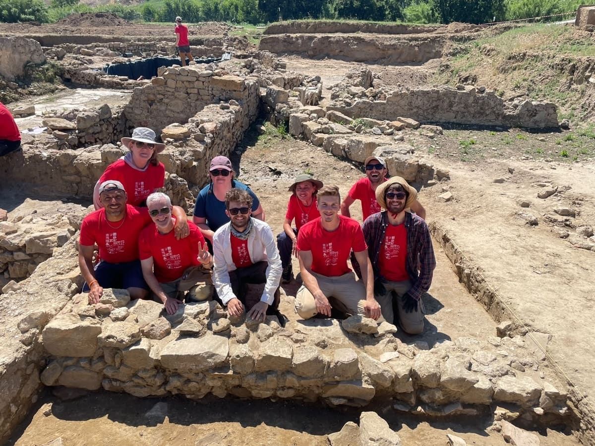 Los arqueólogos excavan un yacimiento con ruinas, muros de piedra, carretillas y una carpa plegable bajo un cielo brillante y soleado.