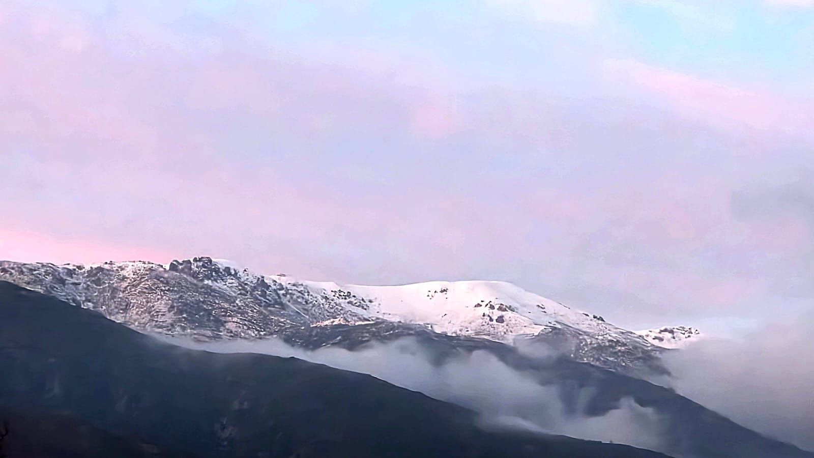 Una cresta montañosa nevada al anochecer, parcialmente oculta por nubes bajas contra un cielo suave de tonos rosados.