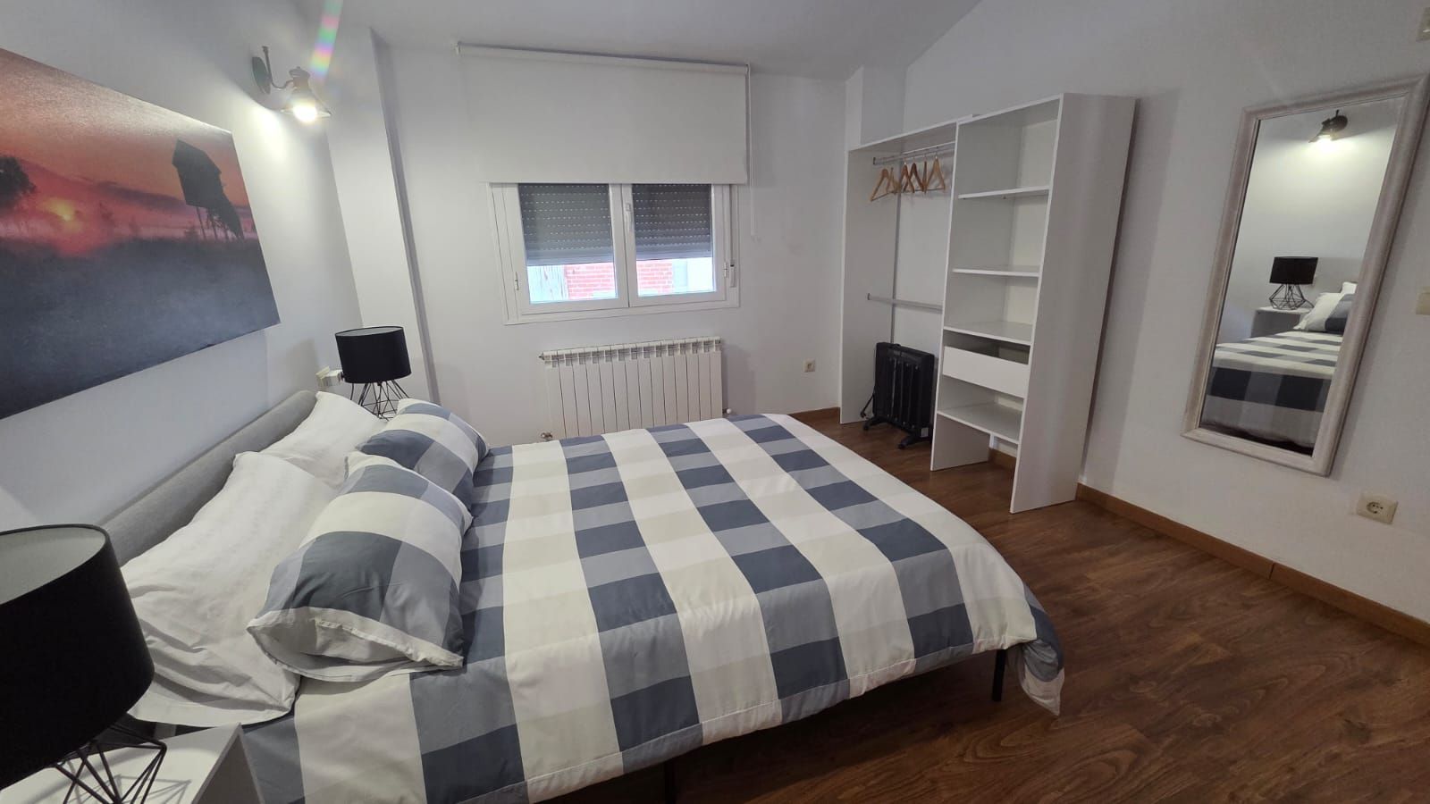 Un dormitorio con una cama con ropa de cama a cuadros azules y blancos, un armario blanco abierto y un gran espejo de pared.