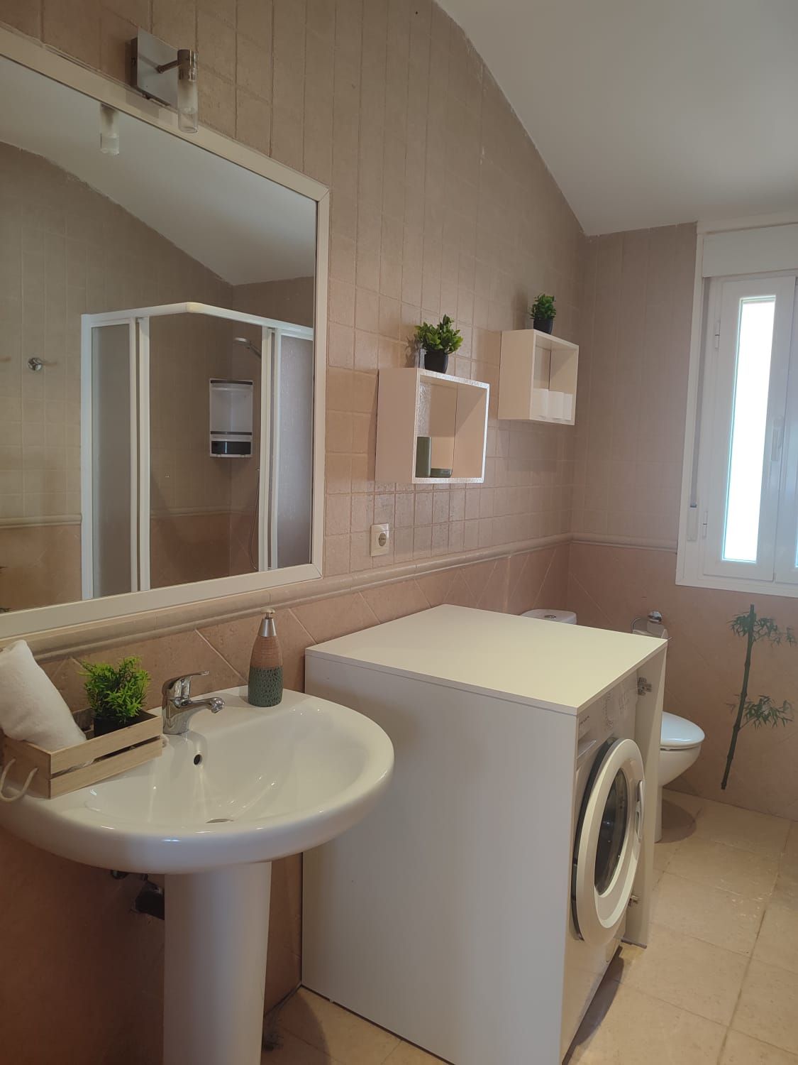 Un baño luminoso con lavabo de pedestal, lavadora blanca, estanterías en la pared y un gran espejo que refleja la ducha.