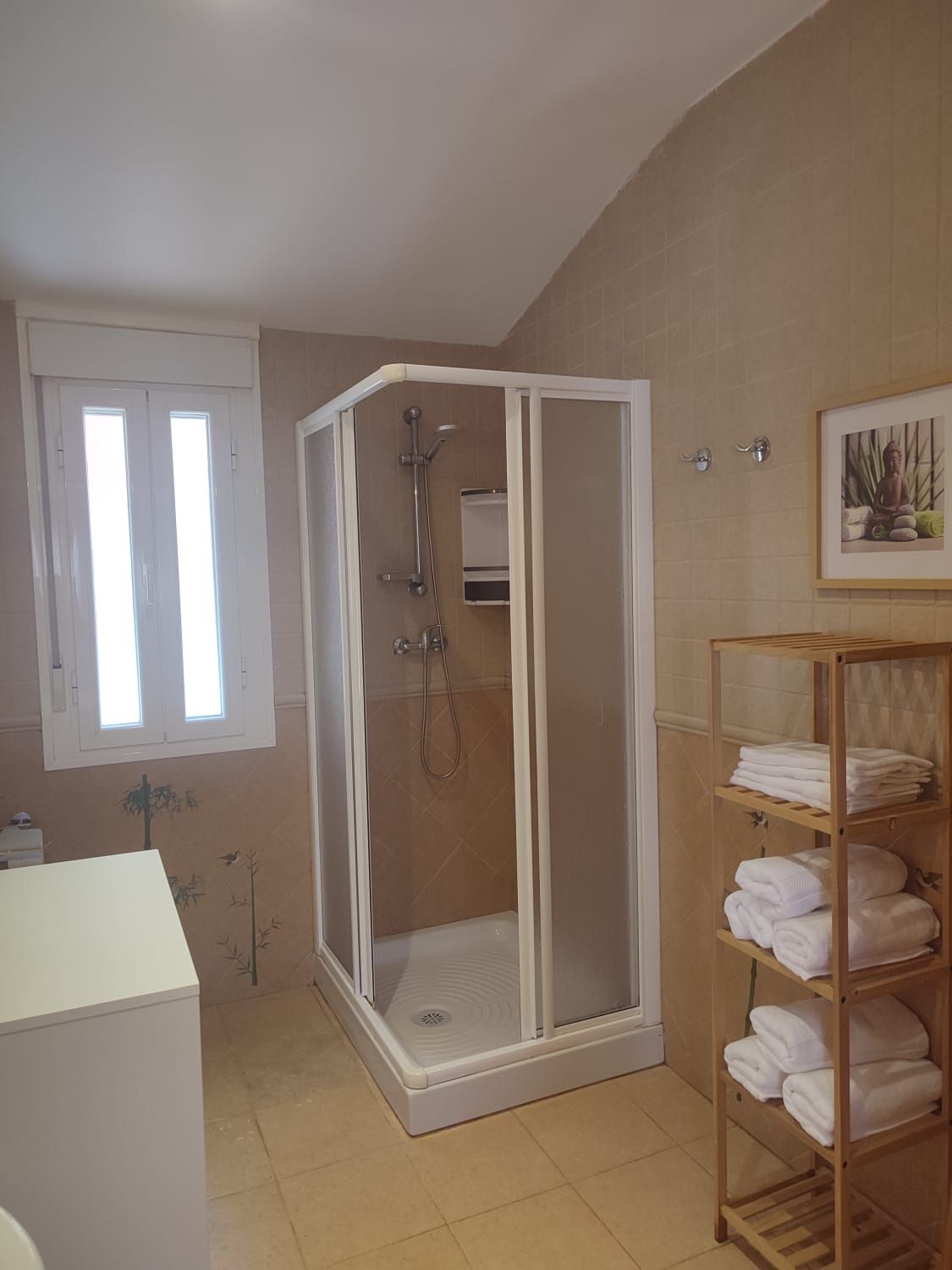 Un baño con una cabina de ducha de esquina blanca, una ventana y una estantería alta de bambú 