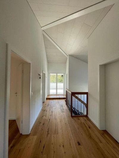 Heller Flur mit Holzboden, weissen Wänden, Dachschräge und einem Treppengeländer in der Nähe eines sonnendurchfluteten Fensters.