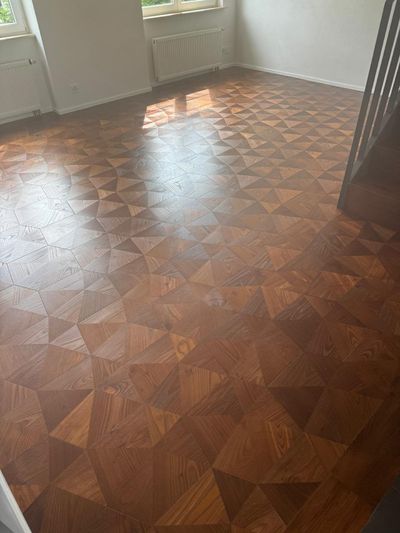 Leeres, sonnendurchflutetes Zimmer mit glänzendem, gemustertem Holzboden und Fenstern entlang der Wand.