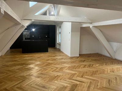 Leeres Dachzimmer mit Fischgrätparkett, weissen Balken und einer dunklen Kücheninsel in Fensternähe.