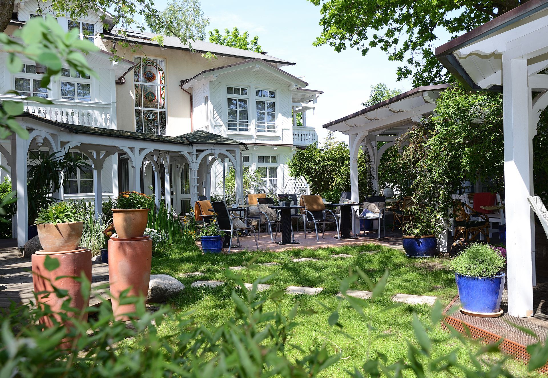 Weiße Villa mit Terrasse, Pavillon und üppigem Garten, sonniger Tag.