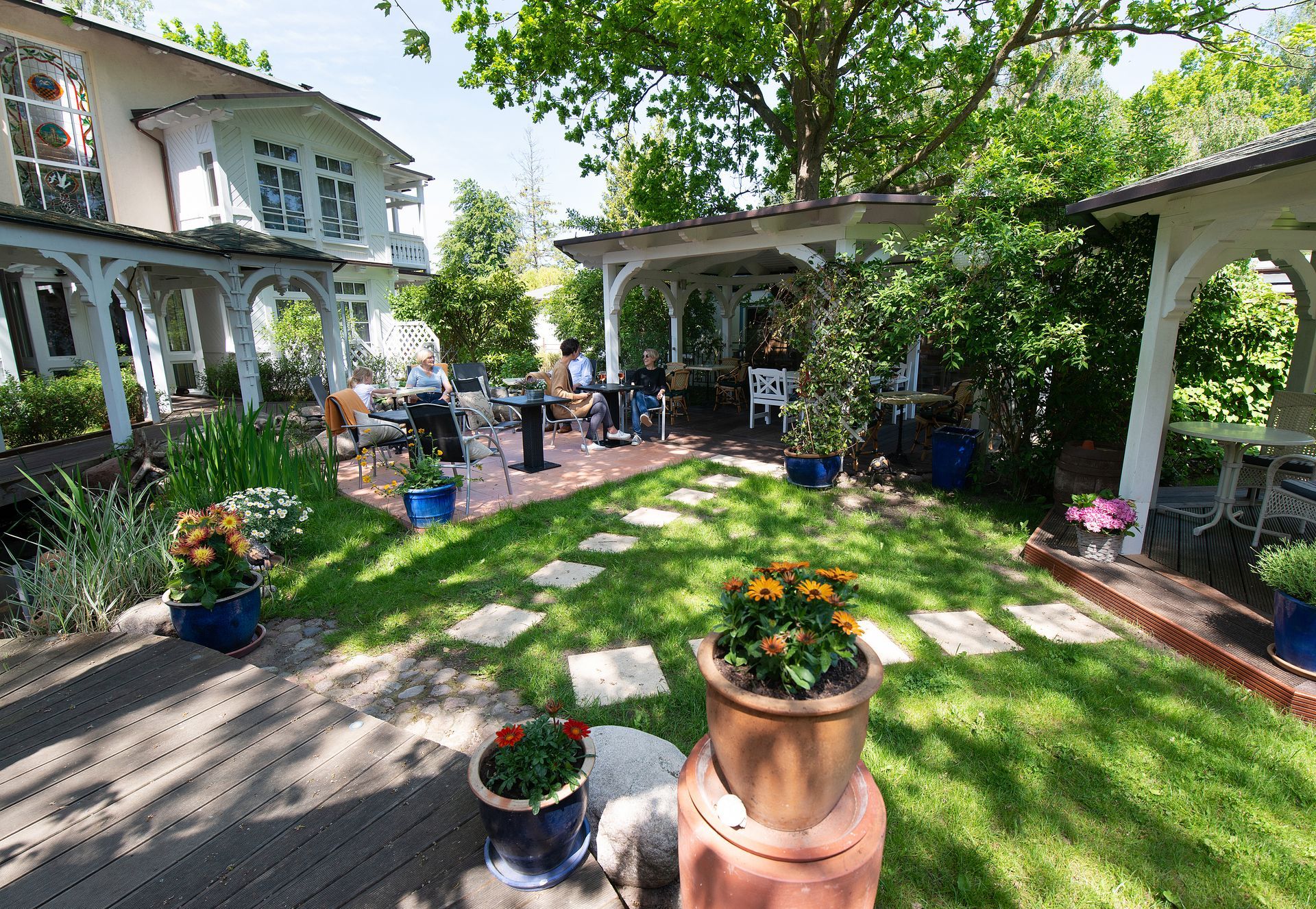 Üppige Außenterrasse mit Pavillons, Tischen, Topfblumen und einem zweistöckigen Haus. Sonniger Tag.