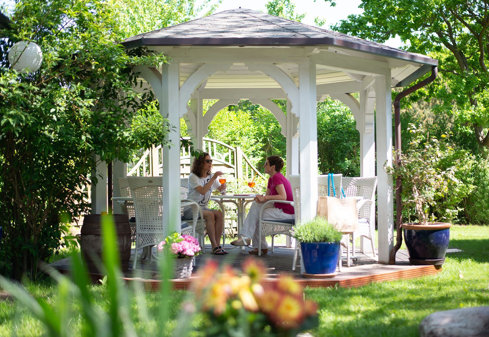 Zwei Frauen in einem weißen Pavillon, trinkend, umgeben von Grün.