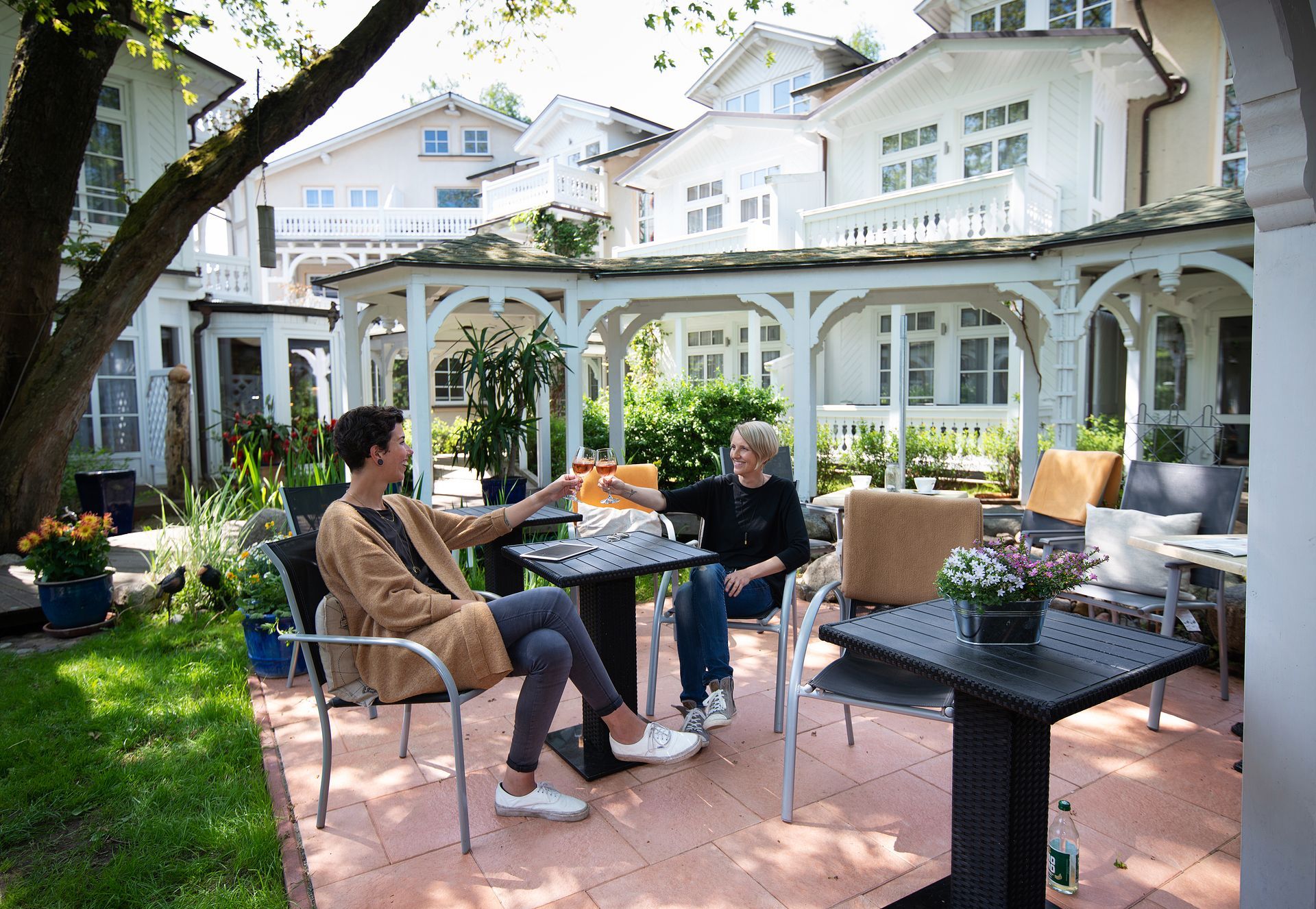 Zwei Personen stoßen in einem Straßencafé mit weißen Gebäuden im Hintergrund mit Getränken an.