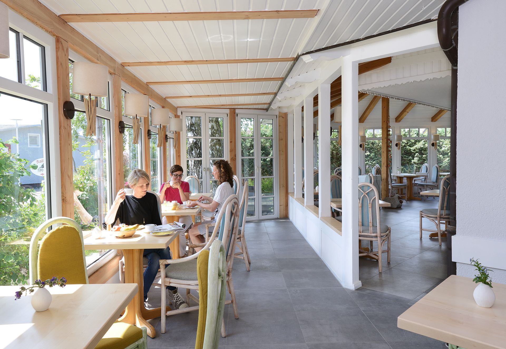 Menschen speisen an Tischen in einem lichtdurchfluteten Wintergartenrestaurant mit großen Fenstern und Holzbalken.