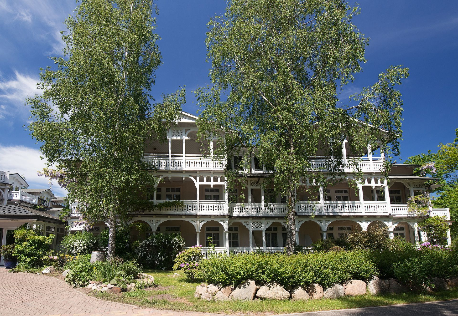 Weiße Holzvilla mit Balkonen, üppigem Grün und blauem Himmel.