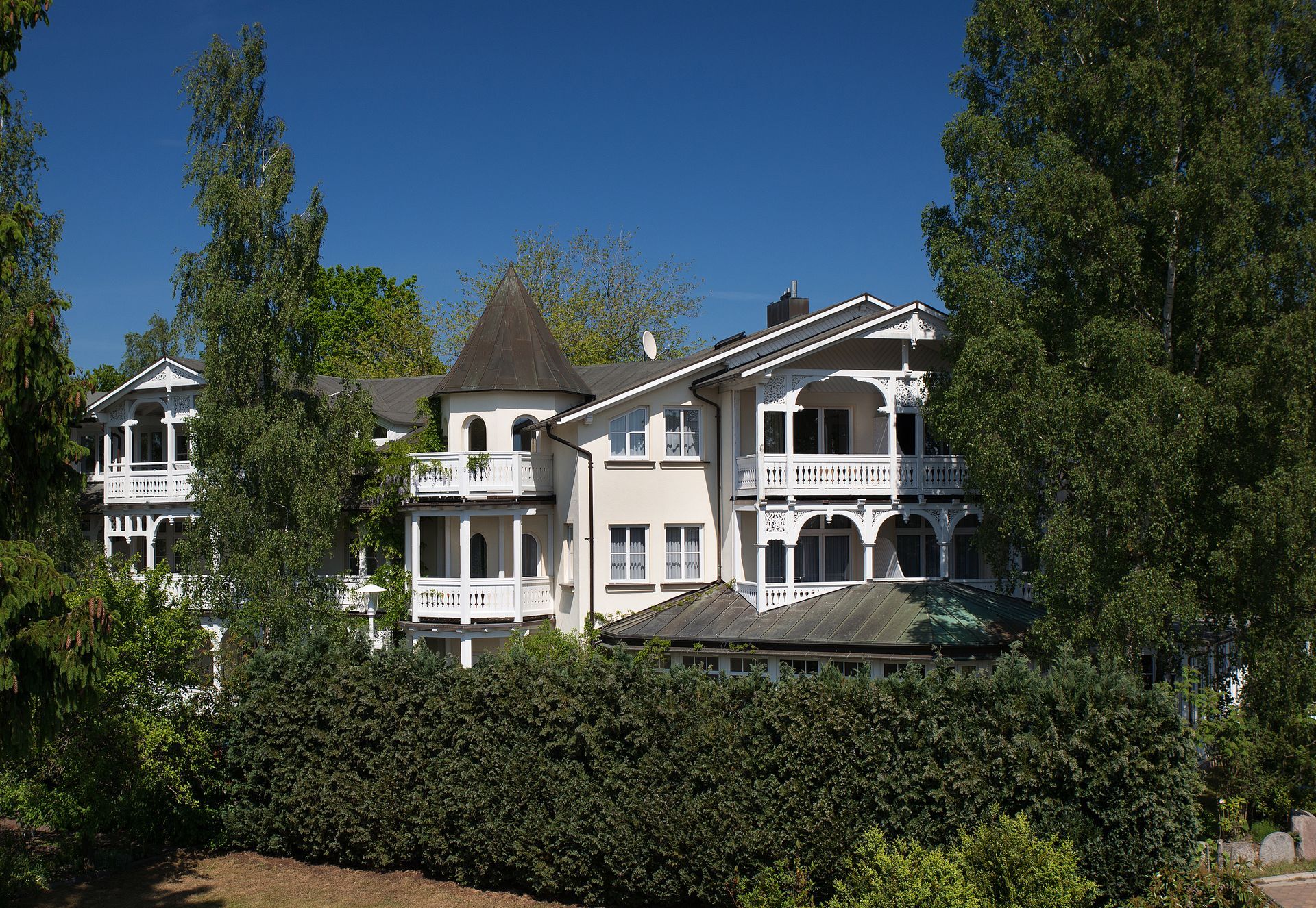 Mehrstöckiges Gebäude in Weiß und Hellbeige mit verzierten Balkonen, grünen Bäumen und einem dunkelblauen Himmel.