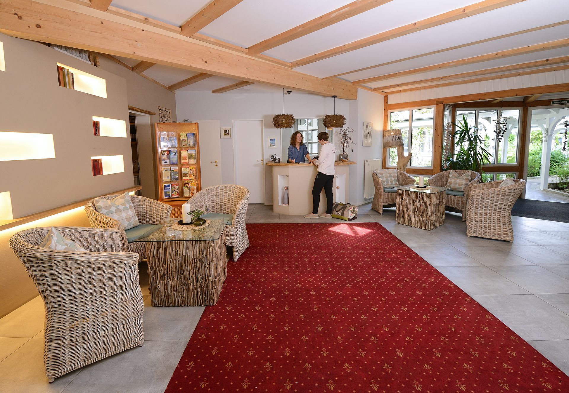 Lobby mit rotem Teppich, Korbmöbeln und Rezeptionstresen, an dem das Personal den Gästen hilft.