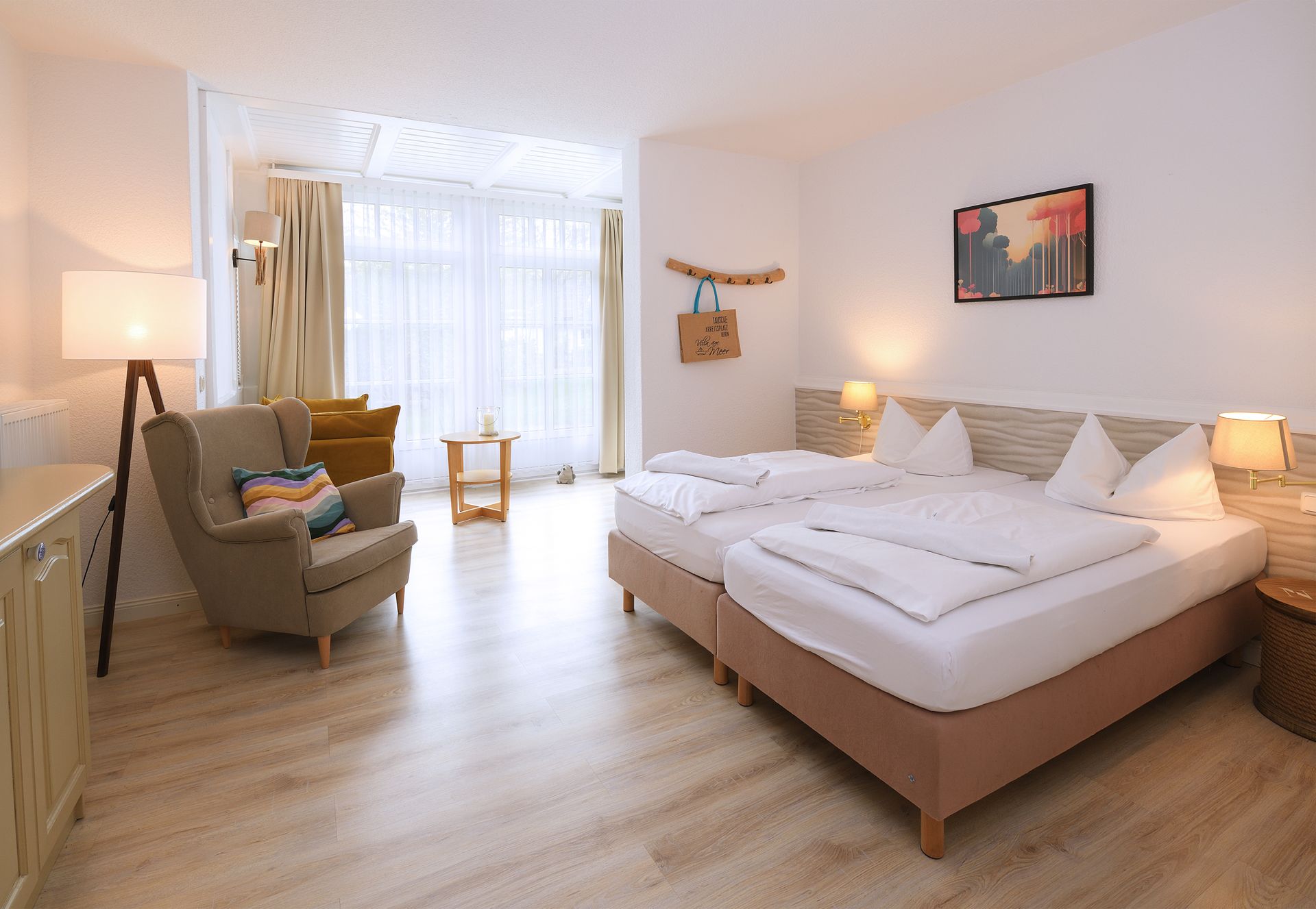 Helles Hotelzimmer mit zwei Einzelbetten, Sessel, Stehlampe und großen Fenstern mit transparenten Vorhängen.
