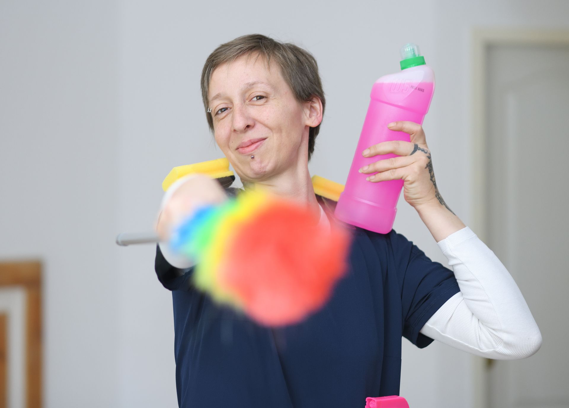Eine Person hält eine rosa Reinigungssprayflasche und streckt einen bunten Staubwedel in Richtung Kamera aus.