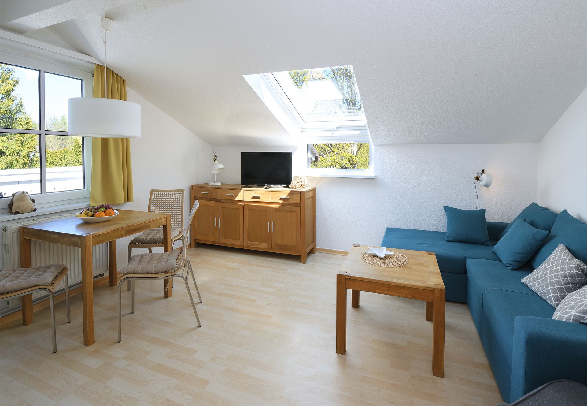 Helles Dachgeschosswohnzimmer mit blauem Sofa, Holztisch, Fernsehschrank, Dachfenstern und großen Fenstern.