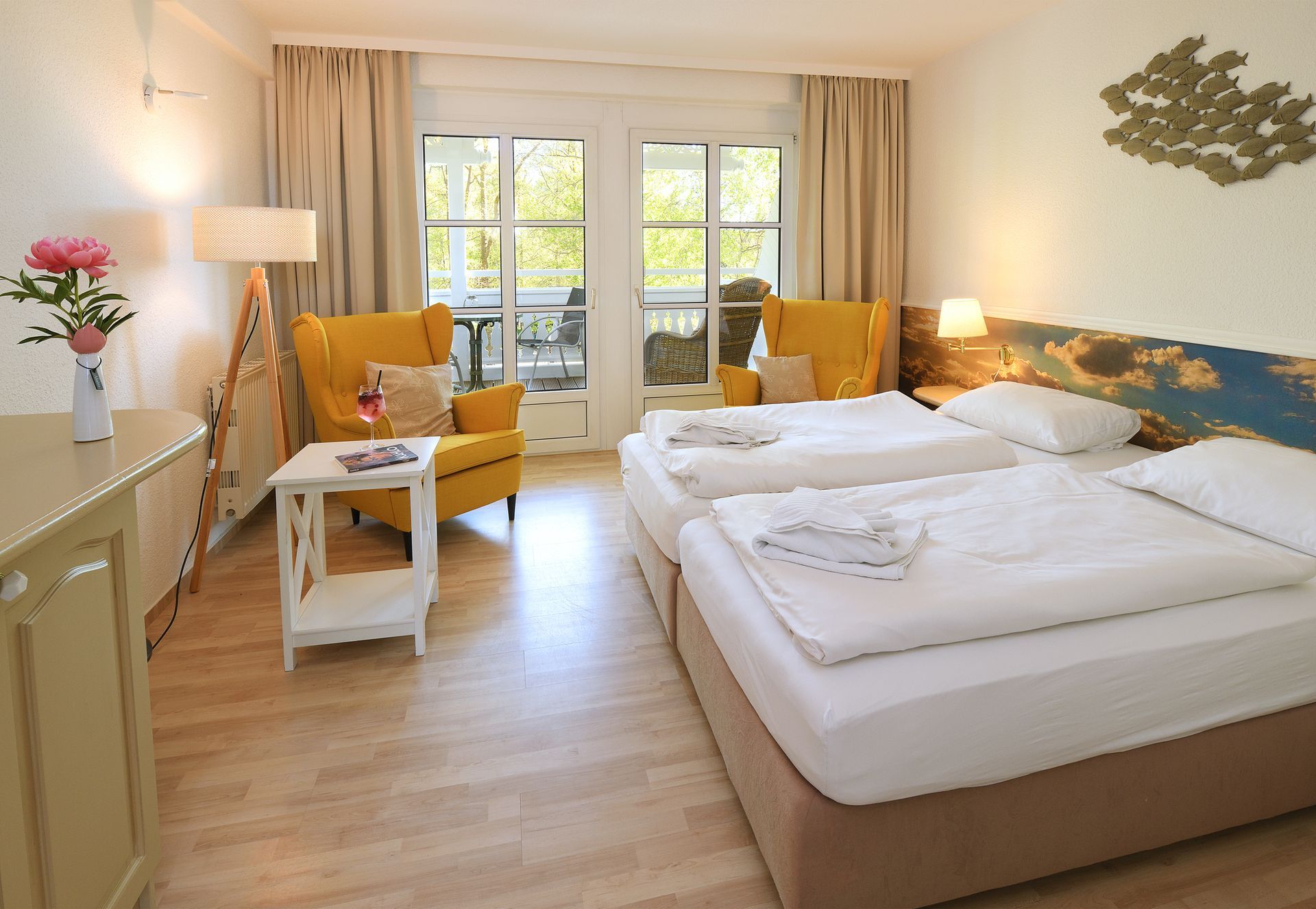 Helles Hotelzimmer mit zwei Betten, gelben Sesseln, großen Fenstern und einem kleinen Tisch am Fenster