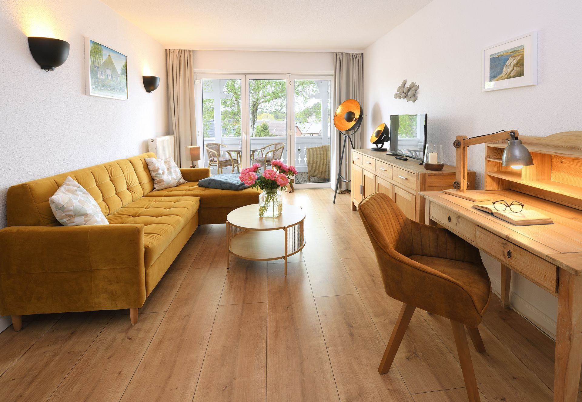 Helles Wohnzimmer mit senffarbenem Sofa, Couchtisch, Schreibtisch und Fenstern mit Blick ins Grüne