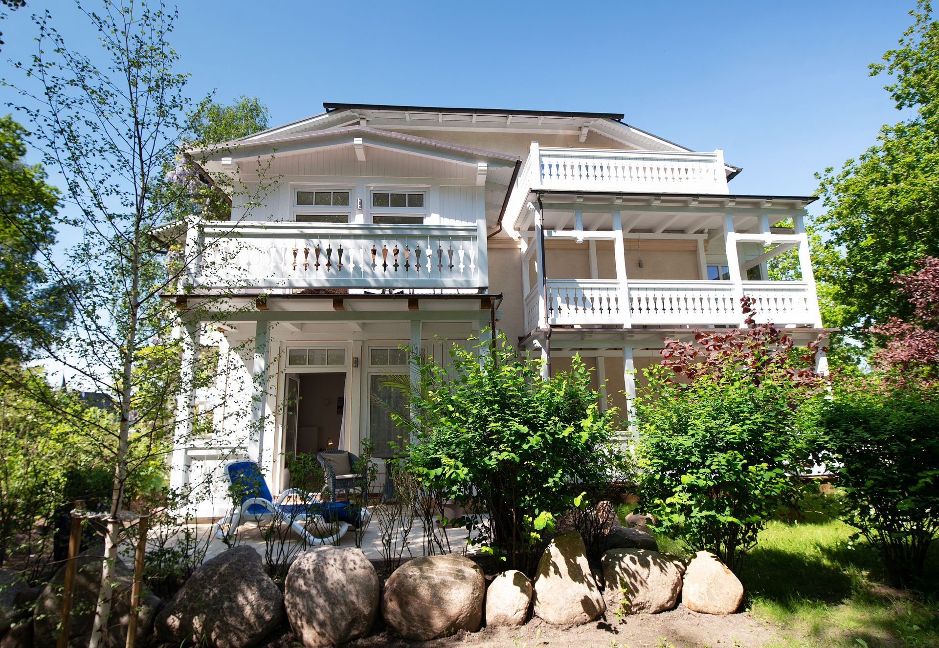 Zweistöckiges weißes Haus mit Balkonen, umgeben von grünen Büschen und Bäumen, vor einem blauen Himmel.