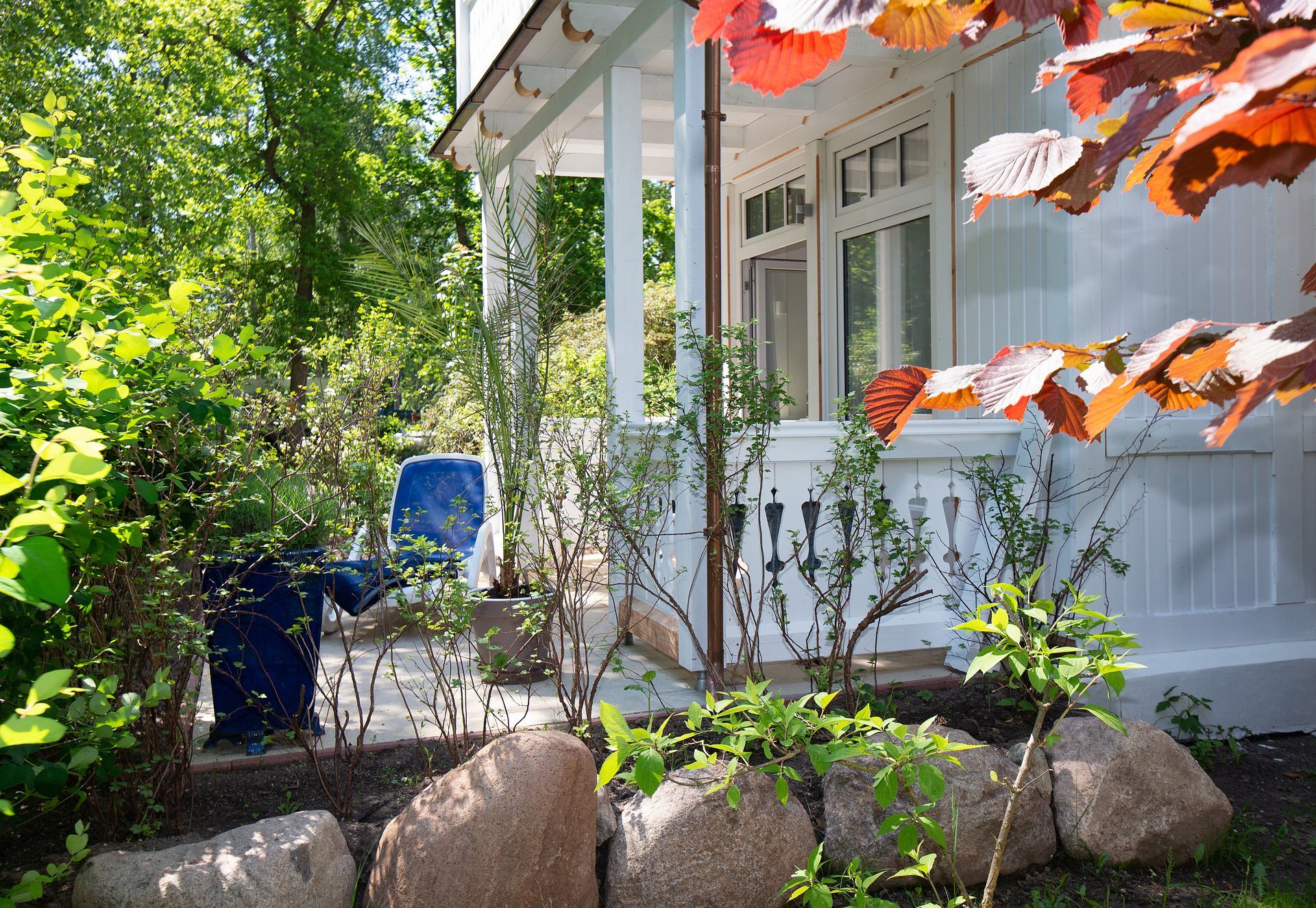 Weiße Veranda mit aufwendiger Holzarbeit, umgeben von üppigem Grün, Steinakzenten und blauen Stühlen.