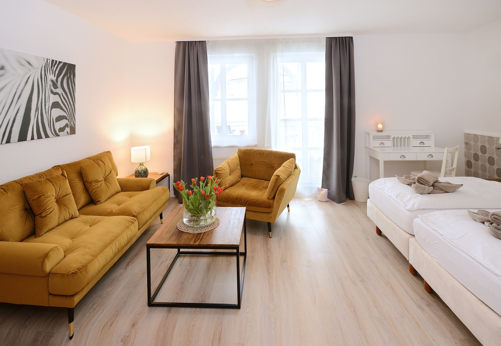 Gemütliches Hotelzimmer mit senffarbenem Sofa, Sessel, Doppelbett, Couchtisch und Schreibtisch.