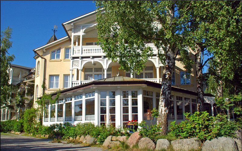 Gelbes Gebäude mit weißen Verzierungen, großen Fenstern und einem Balkon unter blauem Himmel. Bäume und Büsche davor.