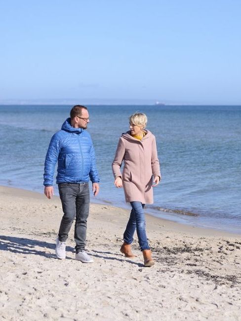 Zwei Personen spazieren an einem Sandstrand; Mann in blauer Jacke, Frau in rosa Mantel, im Hintergrund das Meer.