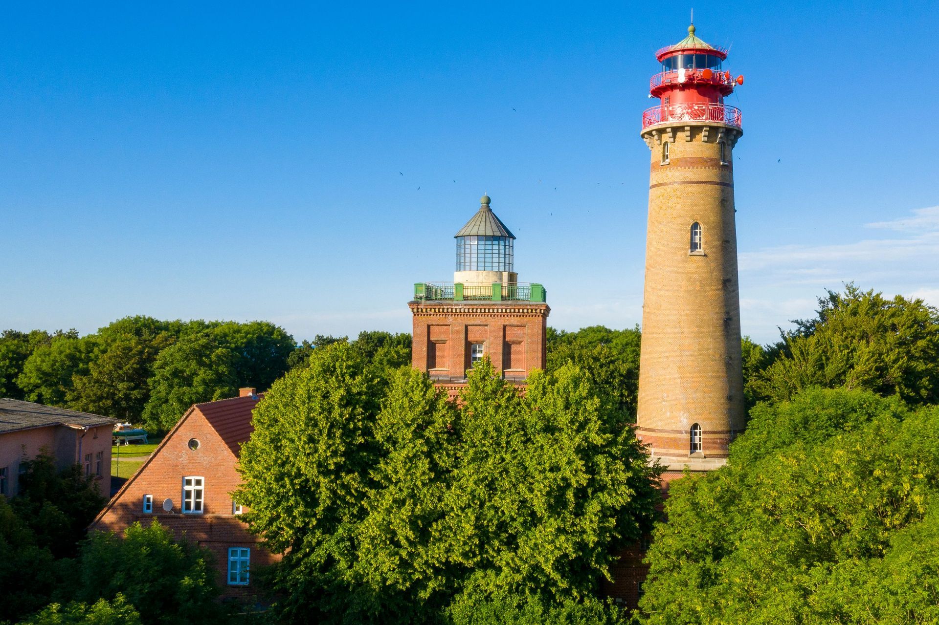 Zwei Leuchttürme, einer hoch und ziegelrot, der andere niedriger, auf Backsteingebäuden, umgeben von Bäumen unter einem blauen Himmel.