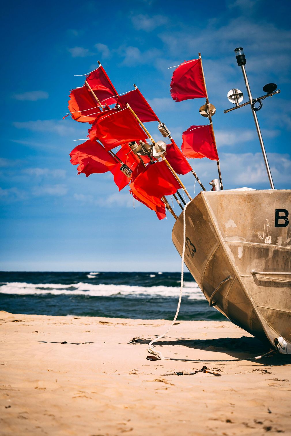 Rote Flaggen auf einem gestrandeten Boot, sandiger Strand, strahlend blauer Himmel und Meereswellen.