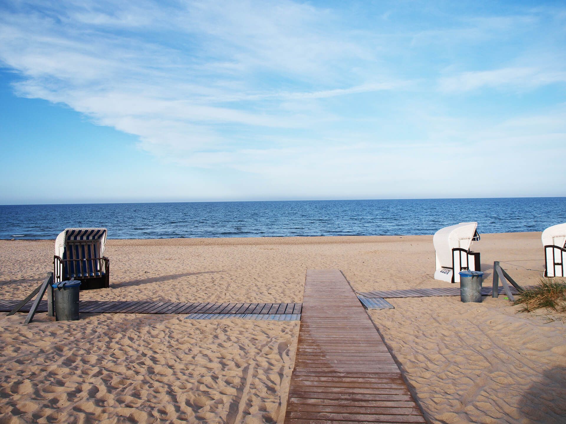 Strand mit Holzsteg, Sand, Meer und Strandkörben unter blauem Himmel.