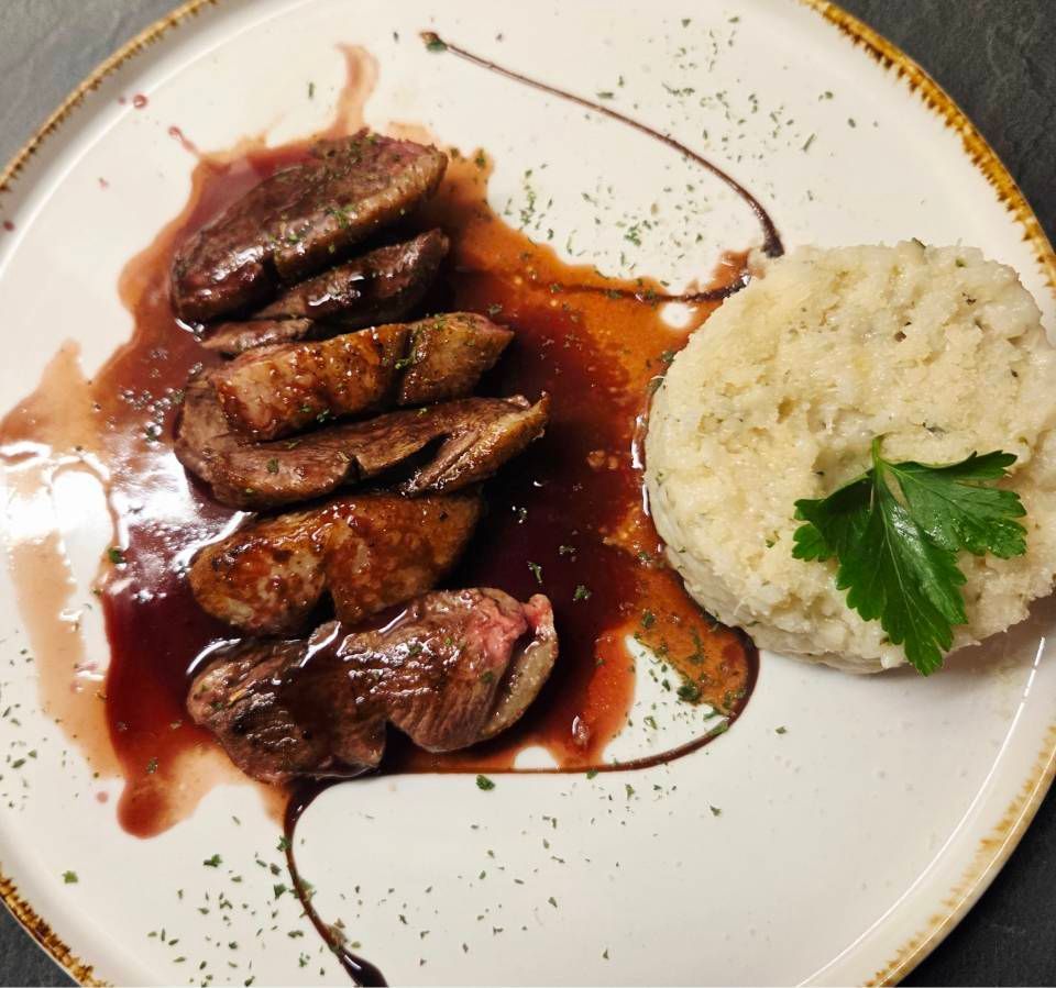 In Scheiben geschnittene, angebratene Entenbrust mit Rotweinsauce und einer Beilage aus cremigem Kartoffelpüree auf einem weissen Teller.