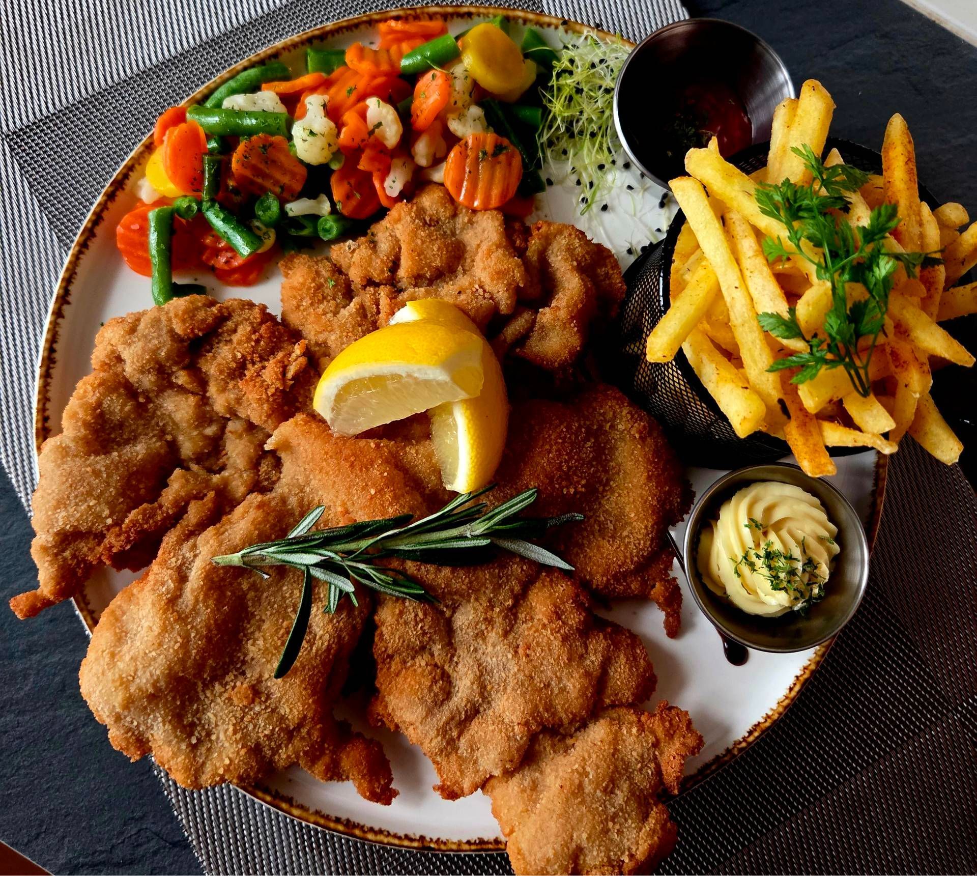 Ein Teller mit paniertem Schnitzel, Pommes frites, sautiertem Gemüse, Zitronenspalten und Dips.
