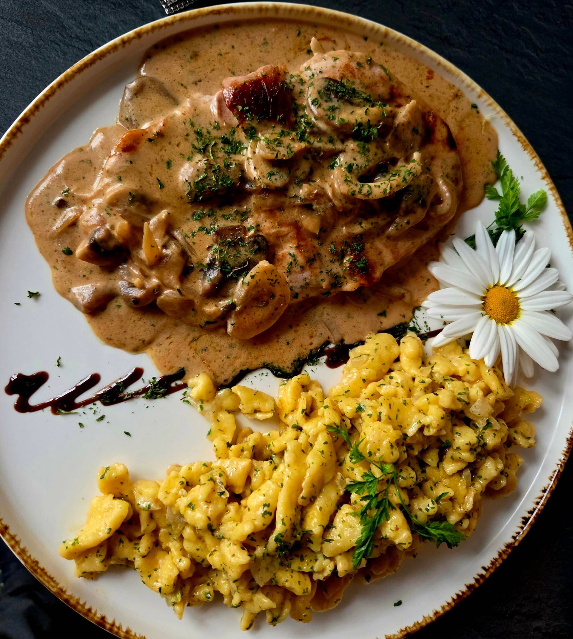Ein Teller Hähnchen in Pilz-Sahnesauce, serviert mit Spätzle, garniert mit Kräutern und einer frischen Gänseblümchenblüte.