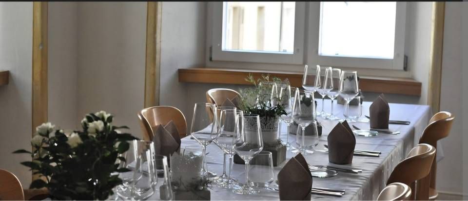 Ein formell gedeckter Esstisch für Gäste mit Weingläsern, weissen Rosen und gefalteten Servietten in einem hellen Raum mit Fenstern.